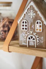 Pepparkakshus för ljus med snökristall, ett pepparkakshus i keramik med vita glasyrdetaljer, står på en trähylla bredvid en burk med kanelstänger och lyser varmt inifrån.