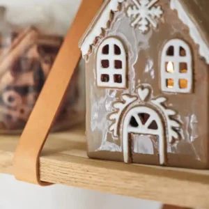 Pepparkakshus för ljus med snökristall, ett pepparkakshus i keramik med vita glasyrdetaljer, står på en trähylla bredvid en burk med kanelstänger och lyser varmt inifrån.