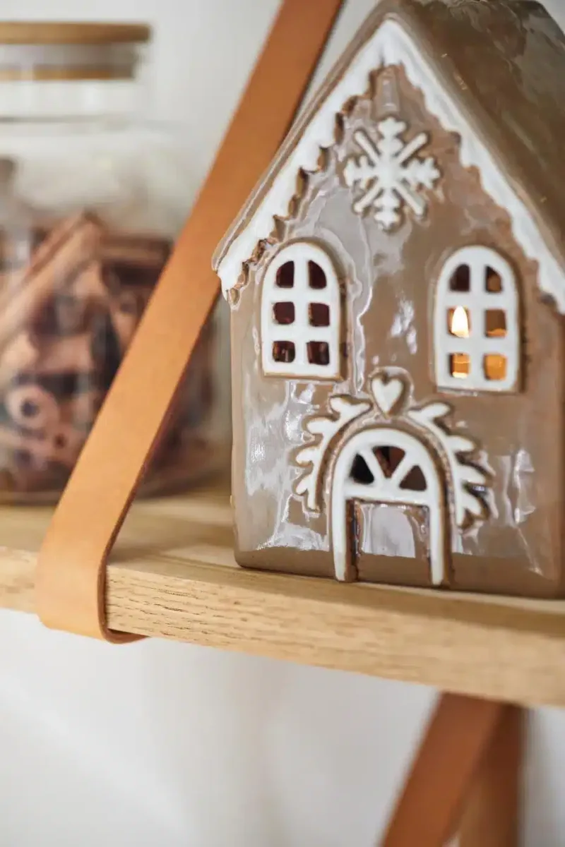 Pepparkakshus för ljus med snökristall, ett pepparkakshus i keramik med vita glasyrdetaljer, står på en trähylla bredvid en burk med kanelstänger och lyser varmt inifrån.