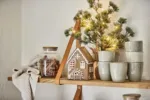 På en trähylla står staplade keramikkoppar, en burk med kanelstänger, en stickad duk, Pepparkakshus för ljus med snökristall och en liten tall med ljusslingor.