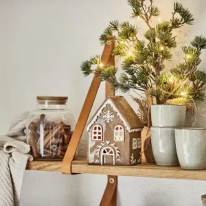 På en trähylla står staplade keramikkoppar, en burk med kanelstänger, en stickad duk, Pepparkakshus för ljus med snökristall och en liten tall med ljusslingor.