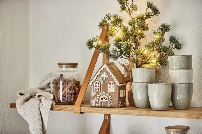 På en trähylla står staplade keramikkoppar, en burk med kanelstänger, en stickad duk, Pepparkakshus för ljus med snökristall och en liten tall med ljusslingor.