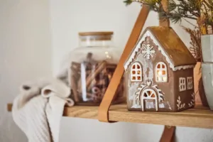 Pepparkakshus för ljus med snökristall, ett dekorativt keramikhus med upplysta fönster, står på en trähylla bredvid en glasburk med kanelstänger, en hopvikt gräddvit stickad duk och en krukväxt med tallgrenar.