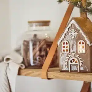 Pepparkakshus för ljus med snökristall, ett dekorativt keramikhus med upplysta fönster, står på en trähylla bredvid en glasburk med kanelstänger, en hopvikt gräddvit stickad duk och en krukväxt med tallgrenar.