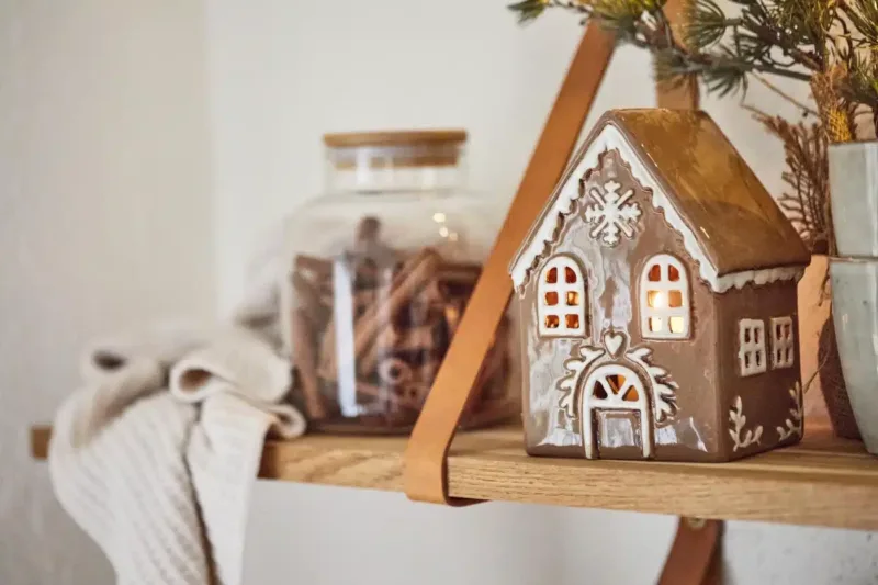 Pepparkakshus för ljus med snökristall, ett dekorativt keramikhus med upplysta fönster, står på en trähylla bredvid en glasburk med kanelstänger, en hopvikt gräddvit stickad duk och en krukväxt med tallgrenar.