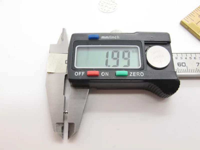 Ett digitalt skjutmått visar 1,99 mm på displayen när det mäter en Silverpärlor runda blanka 2mm/0,9mm pärla, med käftarna öppna och en linjal synlig i bakgrunden.