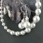 Närbild av Silverpärlor runda blanka 2mm/0,9mm som används i ett pärlhalsband och matchande örhängen med strassdistanser, på en mörk skifferbakgrund bredvid en texturerad snäcka.