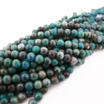 Flera trådar av runda, polerade ädelstenspärlor i blå och gröna nyanser, Krysokoll grön-blå ädelsten 6mm, är buntade med en turkos tofs i ena änden och visas mot en vit bakgrund.