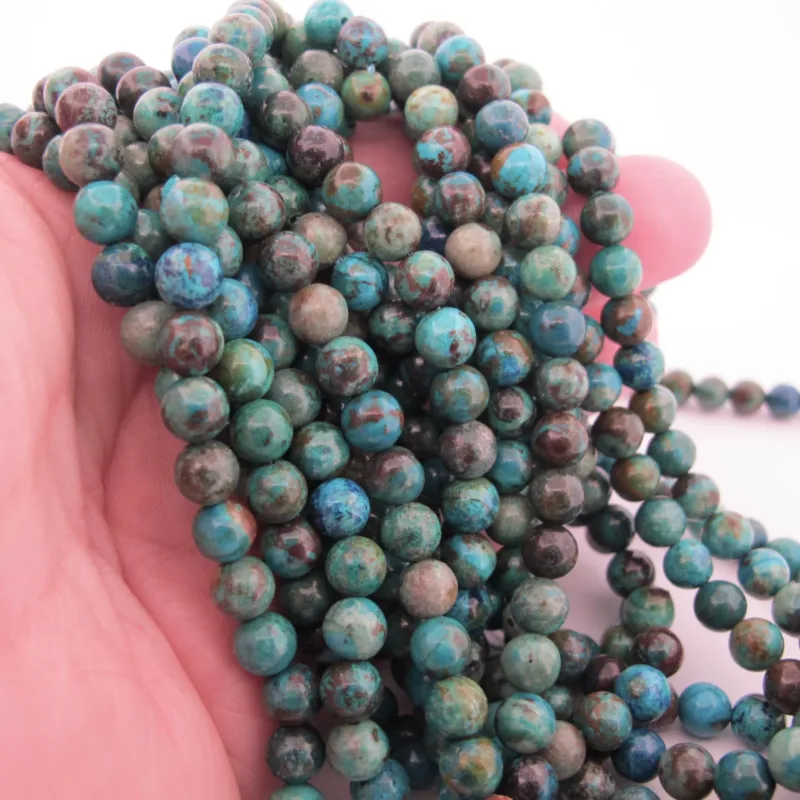 En hand håller i flera trådar av Krysokoll grön-blå ädelsten 6mm runda, polerade pärlor i blå och gröna nyanser mot en vit bakgrund.