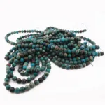 En hög med Krysokoll grön-blå ädelsten 6mm runda pärlor i turkos och mörkgrönt, trädda på blå trådar, ligger löst ringlade och överlappande mot en vit bakgrund.