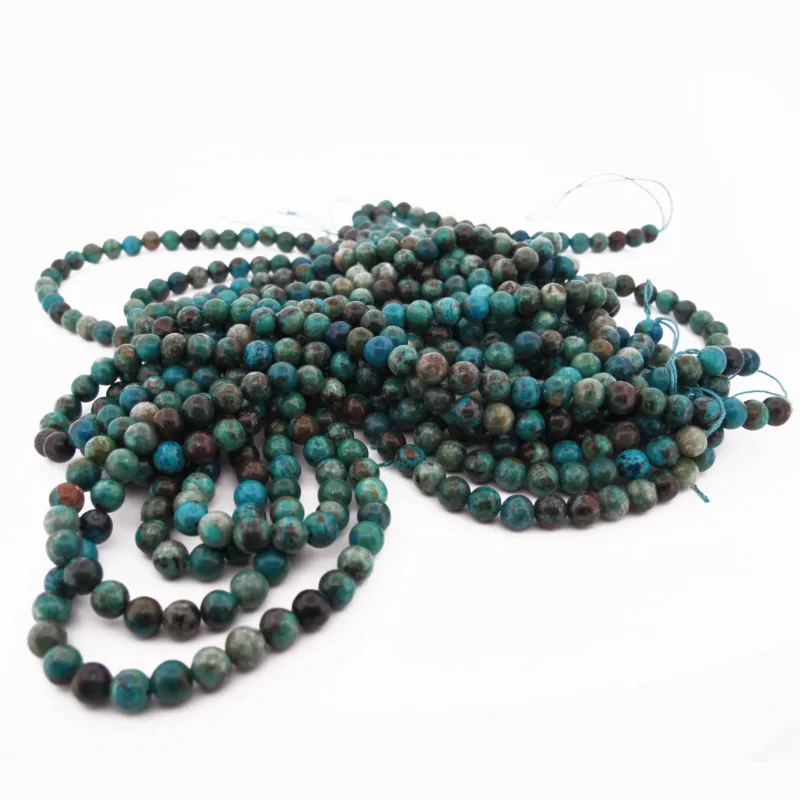 En hög med Krysokoll grön-blå ädelsten 6mm runda pärlor i turkos och mörkgrönt, trädda på blå trådar, ligger löst ringlade och överlappande mot en vit bakgrund.