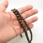 En hand håller i en sträng av Bronsit rund ädelsten 6mm pärlor, runda och mörka med bruna och grönaktiga mönster, mot en enfärgad vit bakgrund.