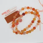 Ett pärlband med Karneol rund ädelsten 4-6 mm matt orange och rosa pärlor ligger bredvid en rulle naturfärgad silkesbrun pärltråd med rödvit etikett. Vissa pärlor är delvis uppträdda på orange tråd.