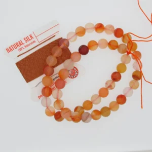 Ett pärlband med Karneol rund ädelsten 4-6 mm matt orange och rosa pärlor ligger bredvid en rulle naturfärgad silkesbrun pärltråd med rödvit etikett. Vissa pärlor är delvis uppträdda på orange tråd.
