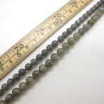 Tre trådar med Labradorit rund ädelsten 6mm pärlor är placerade bredvid en trälinjal markerad i centimeter och millimeter, vilket visar den gradvisa storleksvariationen från små till stora grågröna runda pärlor.