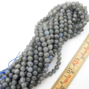 En bunt Labradorit rund ädelsten 6mm pärlor, runda och grå med en blå glans, är uppträdda med blå tråd bredvid en trälinjal markerad i tum och centimeter.