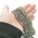 En hand i ett mörkt, ribbat fodral håller flera trådar med 6 mm runda, polerade Labradorit rund ädelstenspärlor, grågröna med en subtil iriserande glans, mot en vit bakgrund.