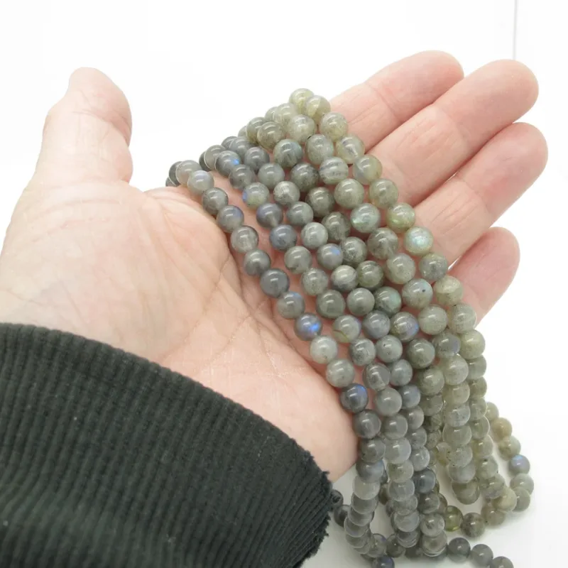 En hand i ett mörkt, ribbat fodral håller flera trådar med 6 mm runda, polerade Labradorit rund ädelstenspärlor, grågröna med en subtil iriserande glans, mot en vit bakgrund.