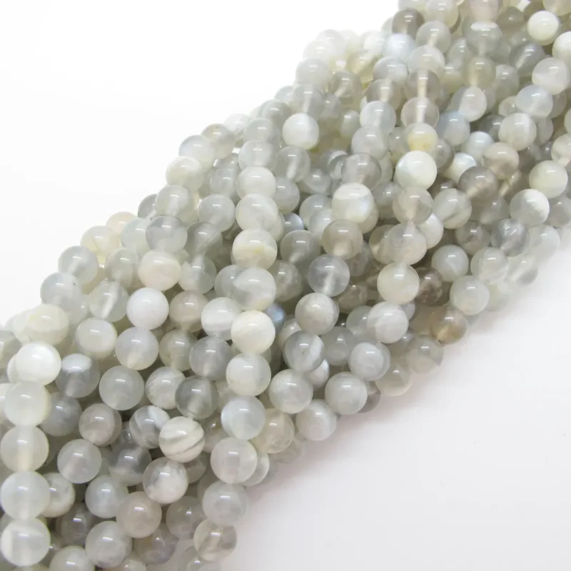 Närbild av flera trådar av Månsten rund ädelsten 6mm, med släta och blanka runda grå och vita pärlor, löst placerade mot en vit bakgrund.