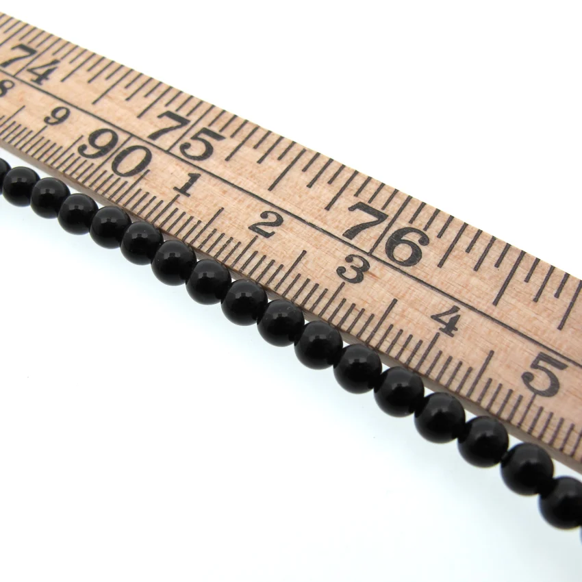 Svart Agat 4mm rund ädelsten 3 En trälinjal markerad i tum och centimeter ligger ovanför en rad små, runda, glänsande svarta pärlor som är arrangerade i en rak linje mot en vit bakgrund.