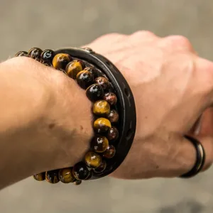 Närbild av en handled som bär en svart ring, ett mörkt läderarmband och Tigeröga rund ädelsten 6mm pärlarmband med bruna och gula stenar mot en suddig grå bakgrund.