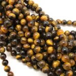 Närbild på Tigeröga rund ädelsten 6mm pärlor med polerade bruna, guldfärgade och gula band, arrangerade i flera trådar mot en vit bakgrund.