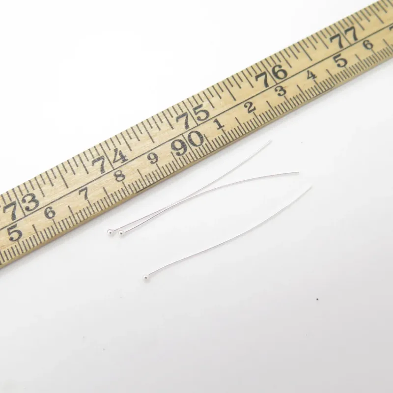 Tre Hattpinne med kula 50mm/0,5mm är placerade bredvid en trälinjal med centimeter- och millimetermarkeringar på en vit bakgrund.