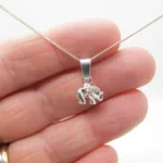 En hand håller i Trekantsögla slät i sterlingsilver, ett slätt halsband i sterlingsilver med ett litet detaljerat elefanthänge, mot en vit bakgrund.