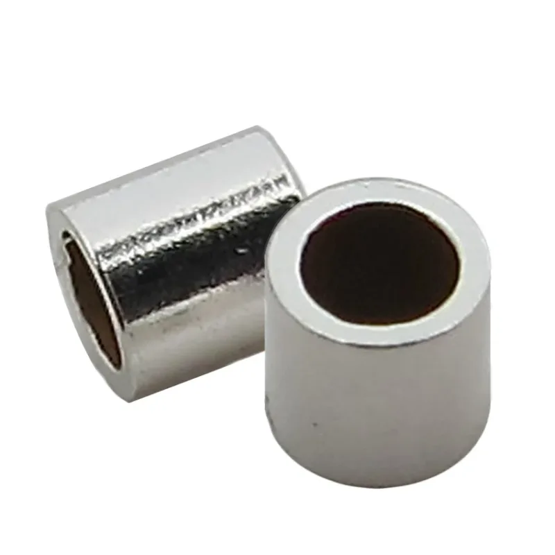 Två små, glänsande Krymphylsa sterlingsilver 2x2mm cylindriska metallpärlor med ihåliga centrum är placerade bredvid varandra på en vit bakgrund.