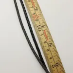 Två flätade lädersnören, ett mörkbrunt och ett svart, är placerade parallellt med en trälinjal på en vit yta. Linjalen visar mått i centimeter.