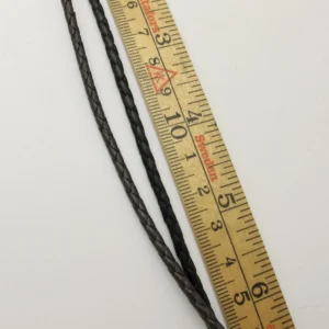 Två flätade lädersnören, ett mörkbrunt och ett svart, är placerade parallellt med en trälinjal på en vit yta. Linjalen visar mått i centimeter.