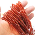 En hand håller i flera trådar av små, runda, polerade rödorange pärlor, med en ljusvit bakgrund som framhäver deras glansiga struktur.