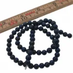 En sträng Lapis Lazuli rund ädelsten 6mm pärlor är upprullad bredvid en trälinjal markerad i centimeter och tum, som visar att de runda, mörkblå stenarna mäter cirka 6 millimeter i diameter.