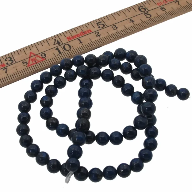 En sträng Lapis Lazuli rund ädelsten 6mm pärlor är upprullad bredvid en trälinjal markerad i centimeter och tum, som visar att de runda, mörkblå stenarna mäter cirka 6 millimeter i diameter.