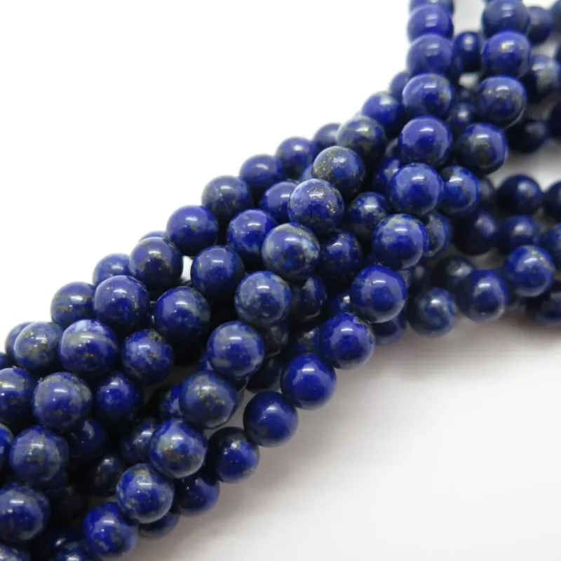 Närbild av flera 6mm runda Lapis Lazuli ädelstenpärlor, polerade mörkblå med subtila guldfläckar, tätt placerade mot en vit bakgrund.