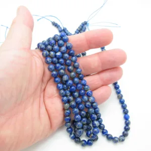 En hand håller flera trådar med Lapis Lazuli rund ädelsten 6mm-runda, polerade blå pärlor med små färgvariationer och några synliga trådändar mot en vit bakgrund.