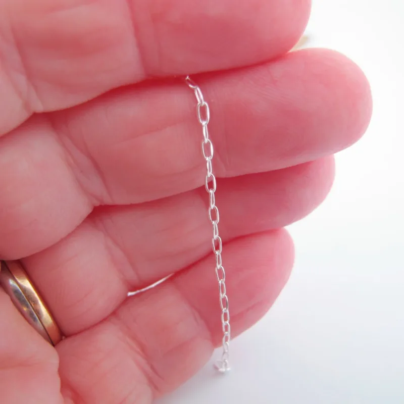 En närbild visar en hand som håller i Silverkedja Oval Facetterad (ca 1,6 mm, 10 cm), en delikat silverkedja med långsträckta ovala länkar. Personen bär en vigselring i guld; den mjukt suddiga bakgrunden är vit.