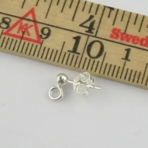 Örstift med kula i Sterlingsilver, en liten silverörhänge med ögla, visas bredvid en trälinjal för skala, vilket indikerar att örhänget är cirka 1 centimeter långt.
