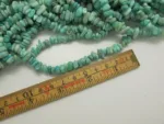 Närbild av Amazonit ädelsten chips (ca 4-6mm) strängar placerade bredvid en centimeterlinjal i trä för skala. De oregelbundna pärlorna har en blågrön nyans.
