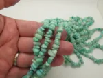 En hand håller i trådar av Amazonit ädelstensflisor ca 4-6 mm, med ljusblågröna oregelbundna pärlor, med ytterligare trådar utspridda på en vit yta i bakgrunden.