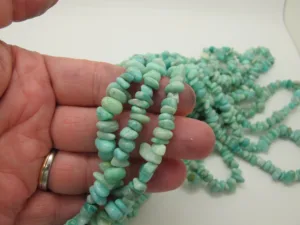 En hand håller i trådar av Amazonit ädelstensflisor ca 4-6 mm, med ljusblågröna oregelbundna pärlor, med ytterligare trådar utspridda på en vit yta i bakgrunden.