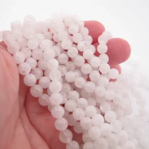 Närbild av en hand som håller i flera trådar av Månsten Vit rund ädelsten 6mm pärlor, med sitt runda, genomskinliga vita utseende mot en vit bakgrund.