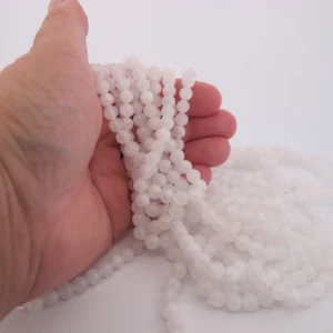 En hand håller flera trådar av Månsten Vit rund ädelsten 6mm pärlor mot en vit bakgrund. De blanka, jämnstora vita ädelstenspärlorna har ytterligare strängar som ligger lösa på ytan.
