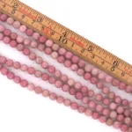 Fyra trådar Rodonit rund smyckesten 4-6mm rosa pärlor är lagda diagonalt under en trälinjal, vilket visar att pärlorna mäter ca 6mm i diameter. Bakgrunden är vit.