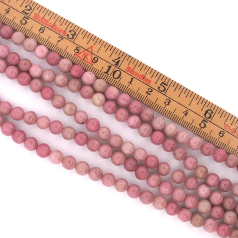 Fyra trådar Rodonit rund smyckesten 4-6mm rosa pärlor är lagda diagonalt under en trälinjal, vilket visar att pärlorna mäter ca 6mm i diameter. Bakgrunden är vit.