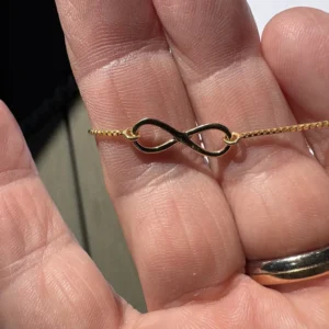 220806327-origpic-30b090 - Fredrix Design & Smyckerian En hand håller i Silverberlock- Evighetstecknet 17.5mm, ett halsband med oändlighetssymbol i guld; personen bär också en guldring på ringfingret.