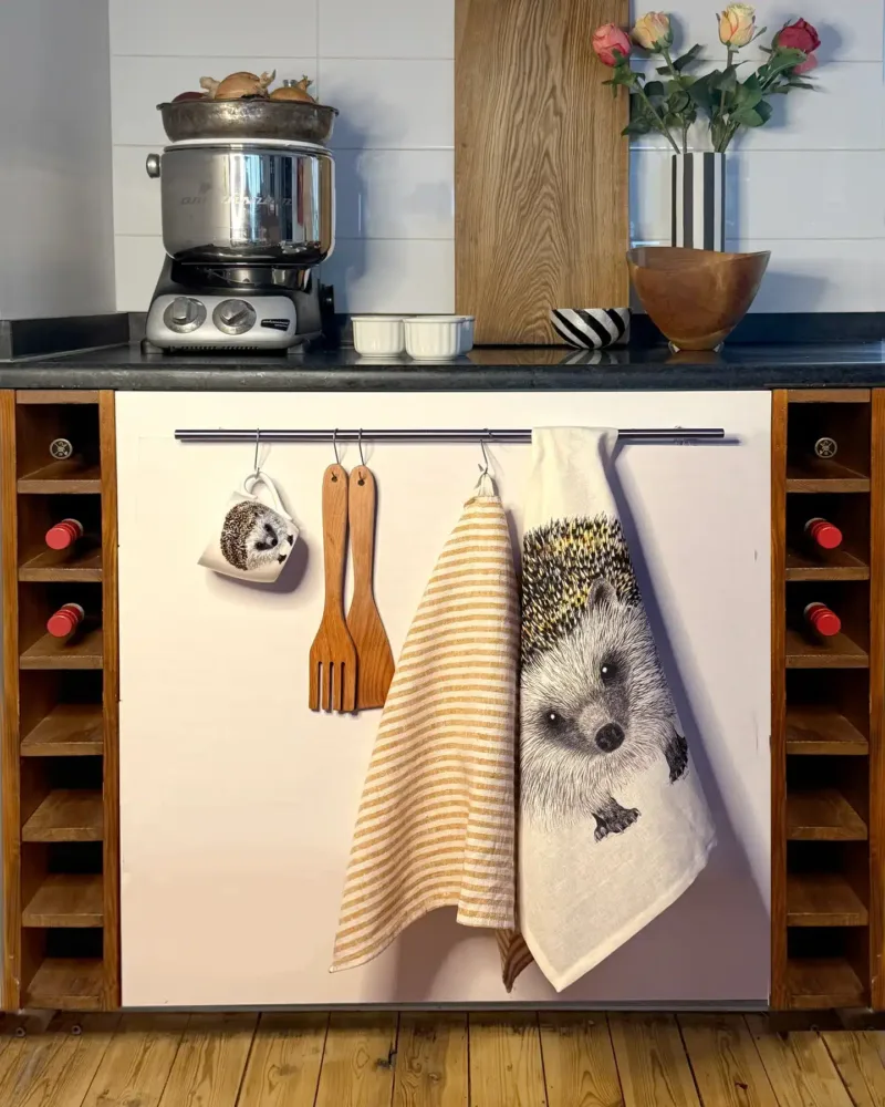 Ett kök har en diskmaskinsdörr dekorerad med Kökshandduk - Igelkott Henry. Träredskap och en randig handduk hänger på en stång, vinflaskor förvaras på var sin sida och en vas med blommor står på bänken.