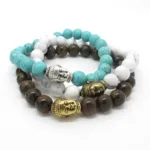 Tre pärlarmband med Buddhahuvudberlocker: ett med turkosa pärlor och en Buddha i silver, ett med vita pärlor och en Buddha i silver samt ett med bruna pärlor och två Buddhahuvuden i guld, alla i en lös stapel.