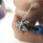 Närbild av en hand som använder en rundnästång för att böja silvertråd till en liten ögla för smyckestillverkning med en Krympkåpa sterlingsilver 3mm, suddig bakgrund.