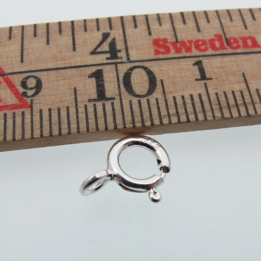 En Ringlås sterlingsilver - öppen ögla 7mm visas nedanför en trälinjal märkt Sverige, som visar centimetermått. Det lilla fjäderringlåset i silver mäter ca 7 mm i diameter.